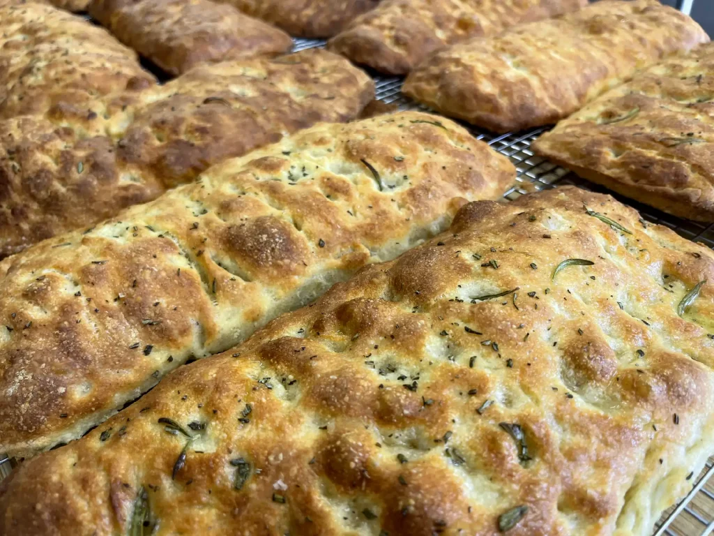 Tray of golden brown herb focaccia loaves cooling on a rack studded with rosemary