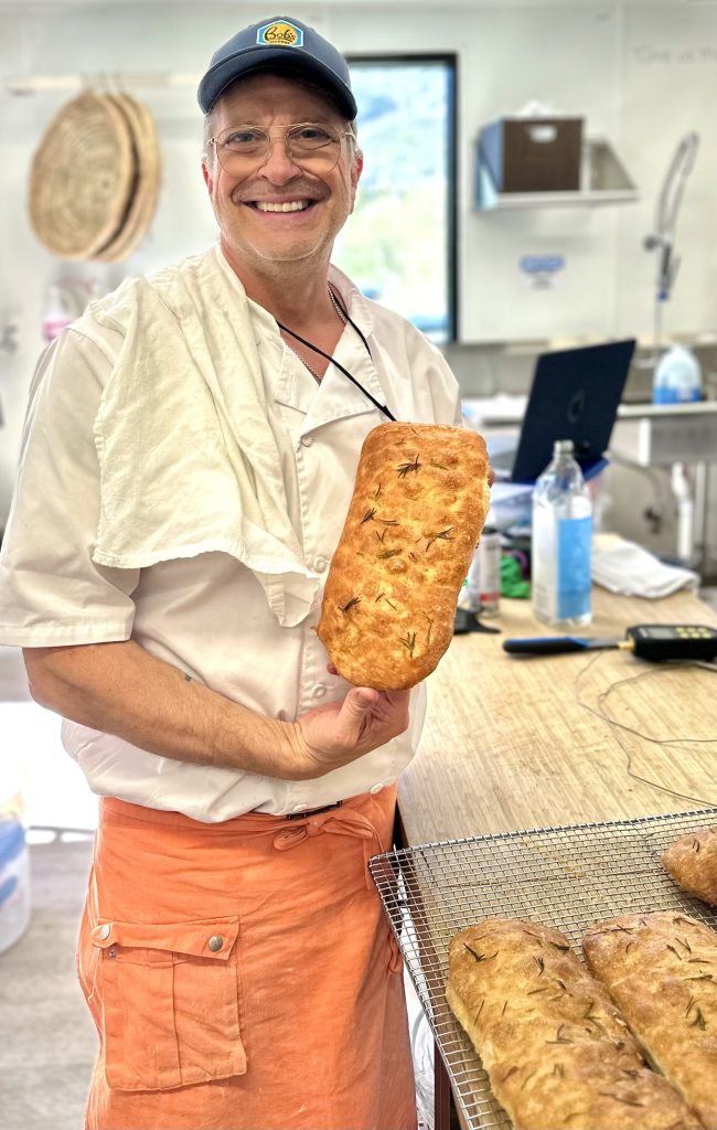 Smiling marvelli owner and baker in a white coat and orange apron holds a long loaf of herb bread in a bakery kitchen