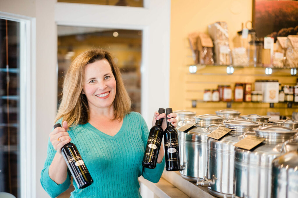 Smiling woman in teal sweater holds bottles of olive oil behind a shop counter with shelves of products in the background