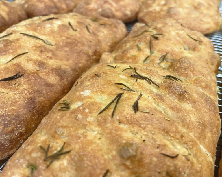 Close up of golden brown marvelli focaccia bread studded with rosemary sprigs on a cooling rack
