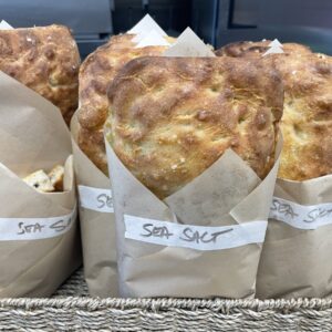 Three large crusty marvelli loaves wrapped in brown paper sleeves labeled sea salt sit in a woven basket on a wooden counter