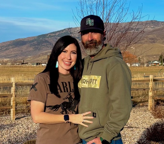 Smiling couple stands arm in arm in a rural field with a wooden fence and mountains in the background woman wearing a brown harley t shirt with turquoise earrings and a watch man in a green hoodie and cap