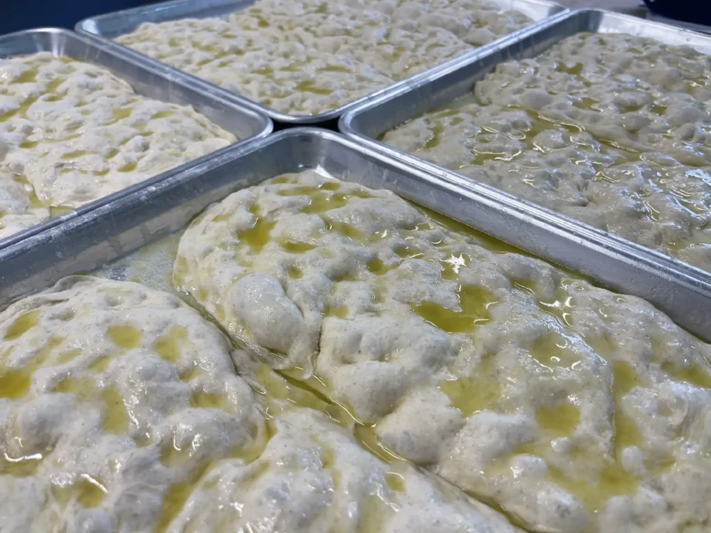 Several metal baking trays filled with risen dough coated with olive oil ready for baking in a bakery