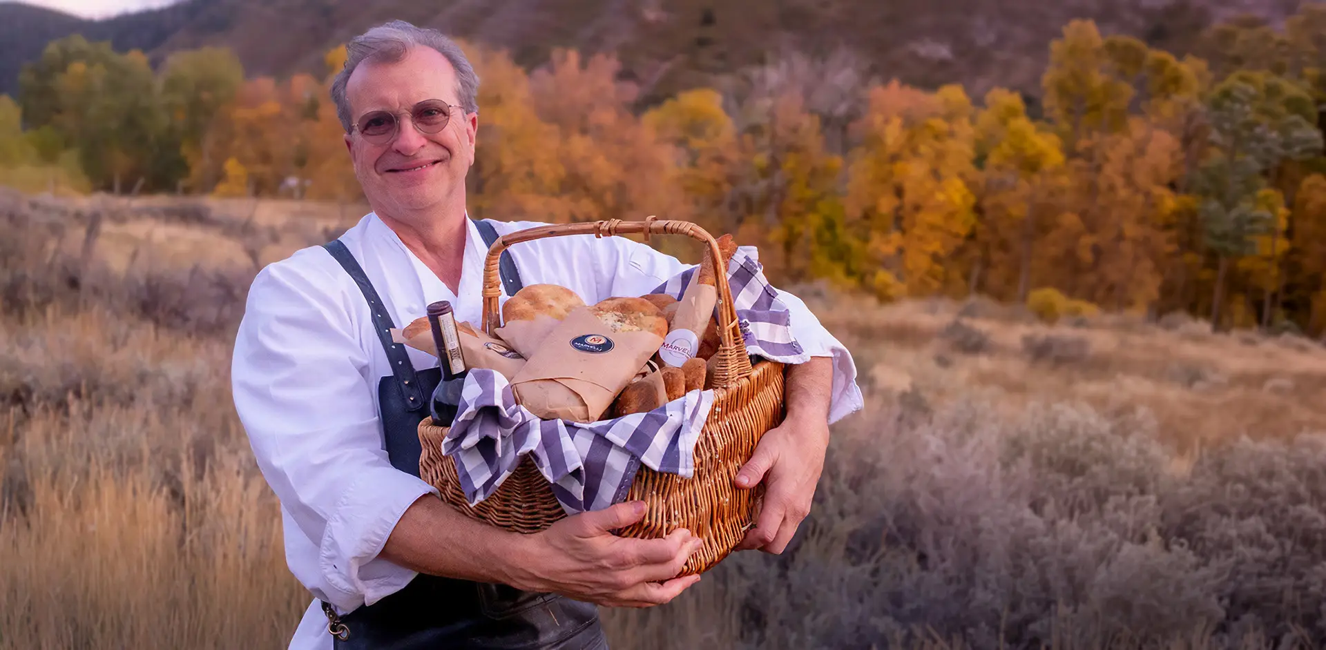 Smiling man in a white shirt and suspenders holding a wicker basket filled with bread outdoors in an autumn landscape