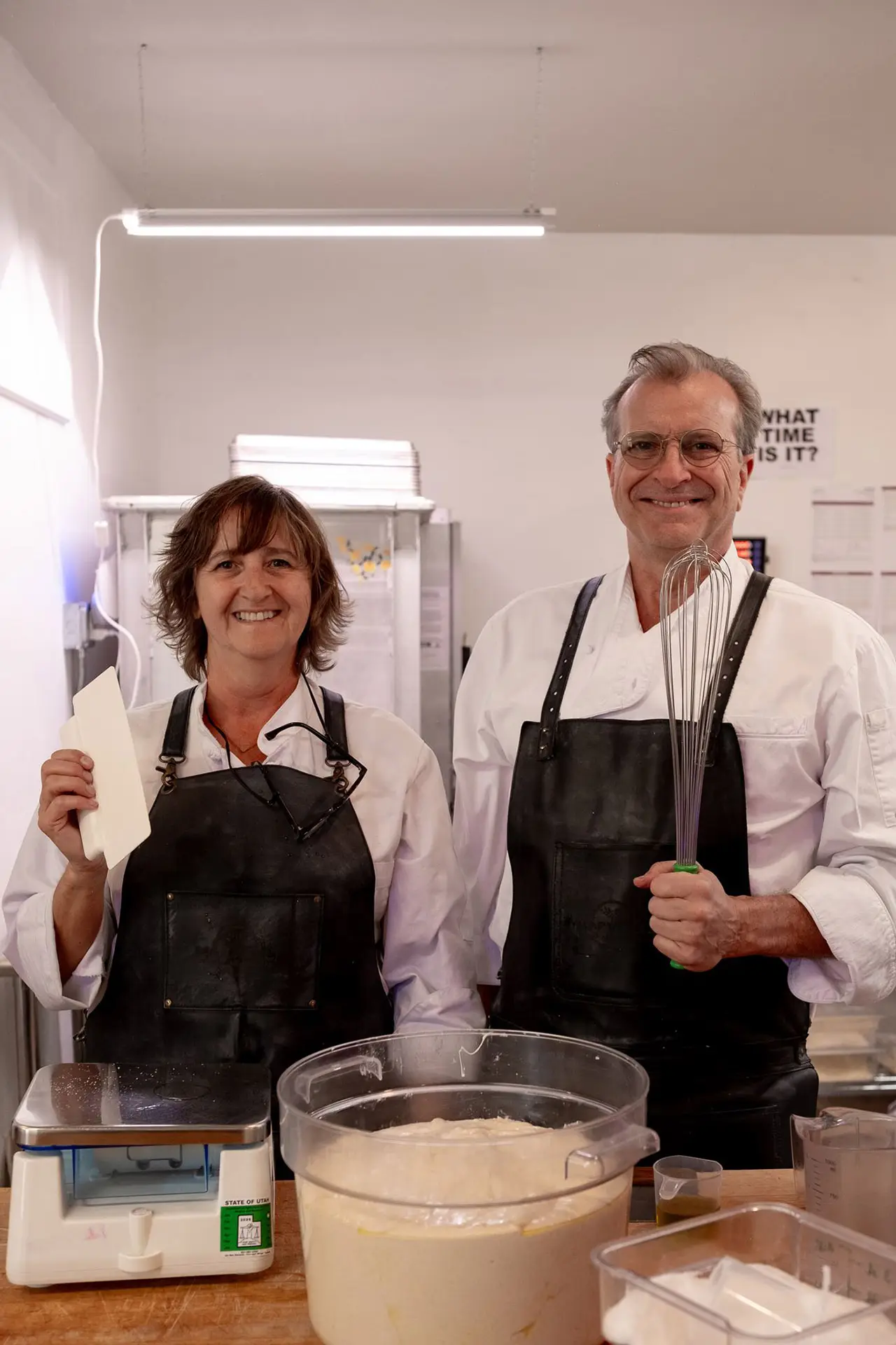 Two bakers in white shirts and black aprons stand at a worktable with a whisk and dairybatter bowls smiling at the camera in a kitchen setting