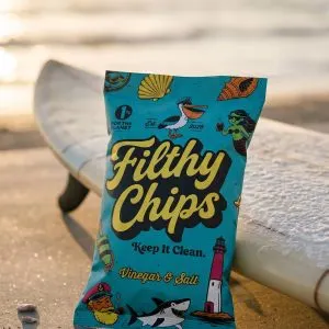 Filthy Chips Vinegar Salt Avocado Oil Kettle Potato Chips 2oz | Marvelli Artisan Breads Bright blue bag of filthy chips on the sand with a surfboard in the background at sunset seaside vibe and beach snack packaging visible